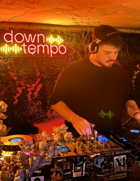 Felipe DJ tocando música en una cabina iluminada con luces cálidas, con un letrero de neón que dice 'downtempo' en el fondo y decoraciones florales