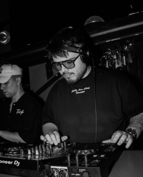 Felipe DJ tocando música con una persona al lado. Felipe, tiene puesto unos lentes y una remera negra.